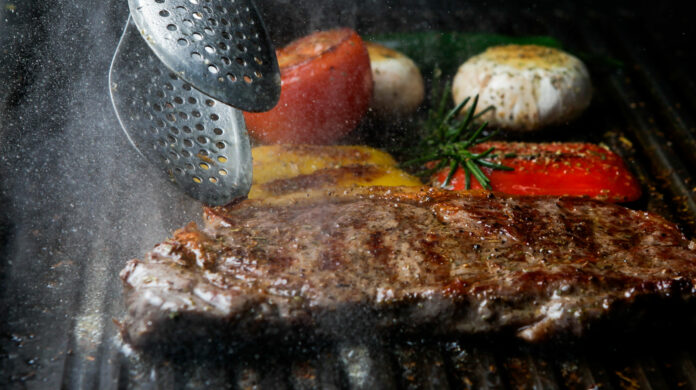Side view grilled steak with rosemary and mushrooms and barbecue tongs in barbecue on dark background